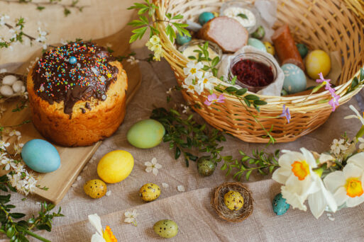 Homemade easter bread, natural dyed easter eggs, ham, beets, butter in basket on rustic table with spring blossoms and linen napkin. Top view. Traditional Easter food