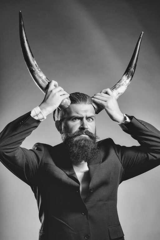 Handsome young man with long beard and moustache in black jacket holding animal antlers in studio on grey background
Мужчина Овен